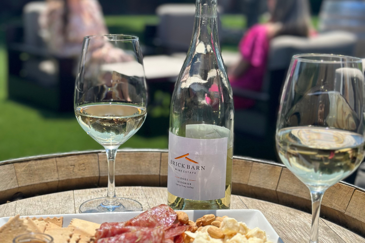 Two glasses and a bottle of Brick Barn white wine with a charcuterie board on a barrel with two women chatting in the back ground