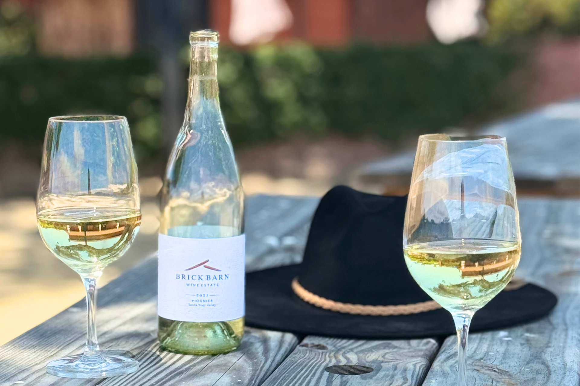A bottle of Brick Barn wine and glasses sitting on a wood picnic bench with a black hat